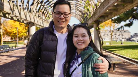 A man stands with his arm around a woman's shoulder under a wooden arch in a public park on a sunny day