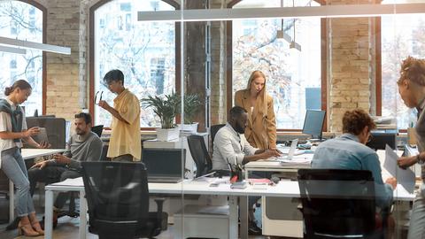 People work alone and in small groups at desks arranged around an office with large windows and exposed brick walls