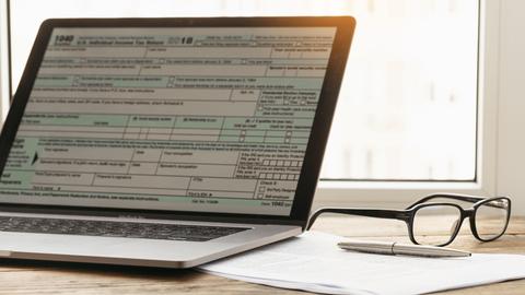A laptop with a tax form displayed on the screen next to a pair of glasses, some papers, and a pen arranged on a desk in front of a window