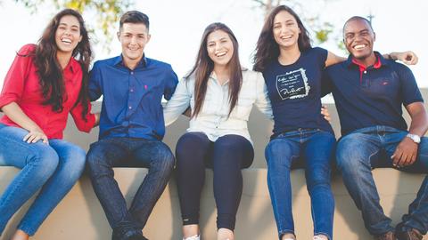Five smiling students sit on a wall outside