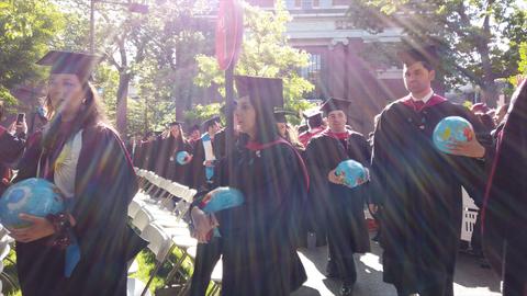 Graduates in caps and gowns process into Harvard Yard under bright sunshine