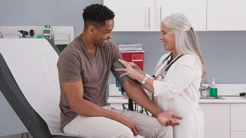 A doctor in a white coat straps a blood pressure cuff around a patient's arm in a doctor's office