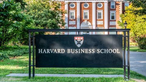 A sign reading "Harvard Business School" on a grassy lawn in front of a red brick building with white columns