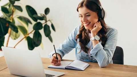 A woman smiles and takes notes while watching a webinar on a laptop