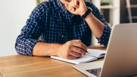 A man takes notes in a notebook while watching a webinar on a laptop