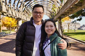 A man stands with his arm around a woman's shoulder under a wooden arch in a public park on a sunny day