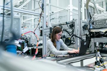 A woman with glasses works at a laptop in an engineering lab space