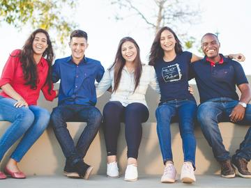Five smiling students sit on a wall outside