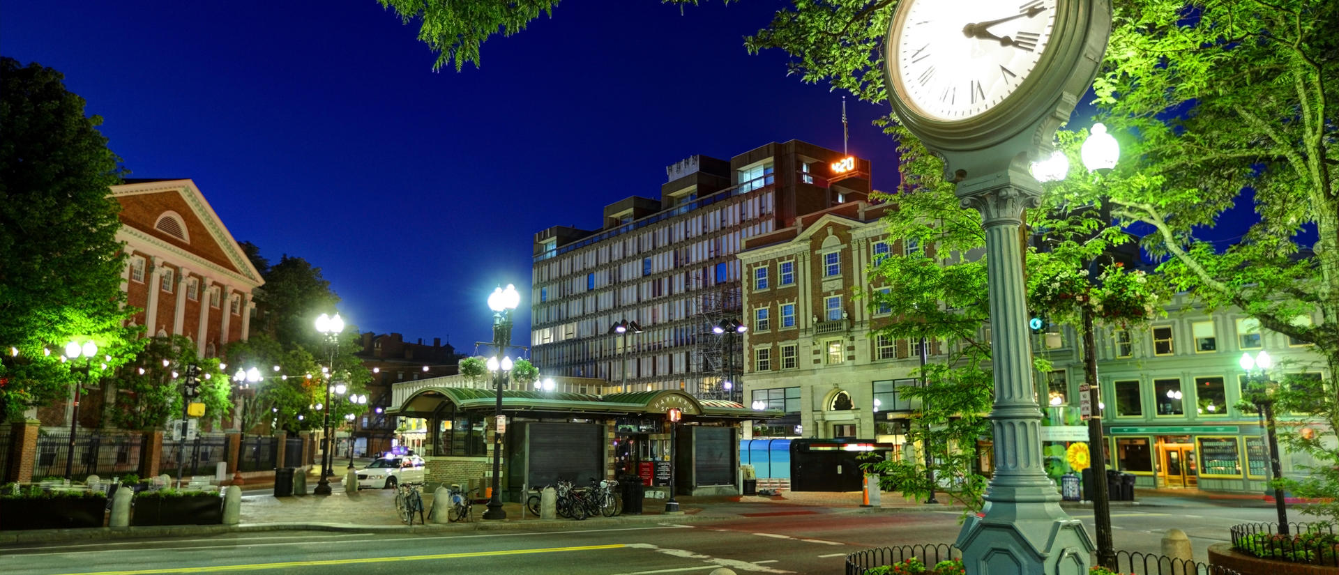 A city intersection at night with an illuminated clock in the foreground and a mix of historic and newer buildings in the background
