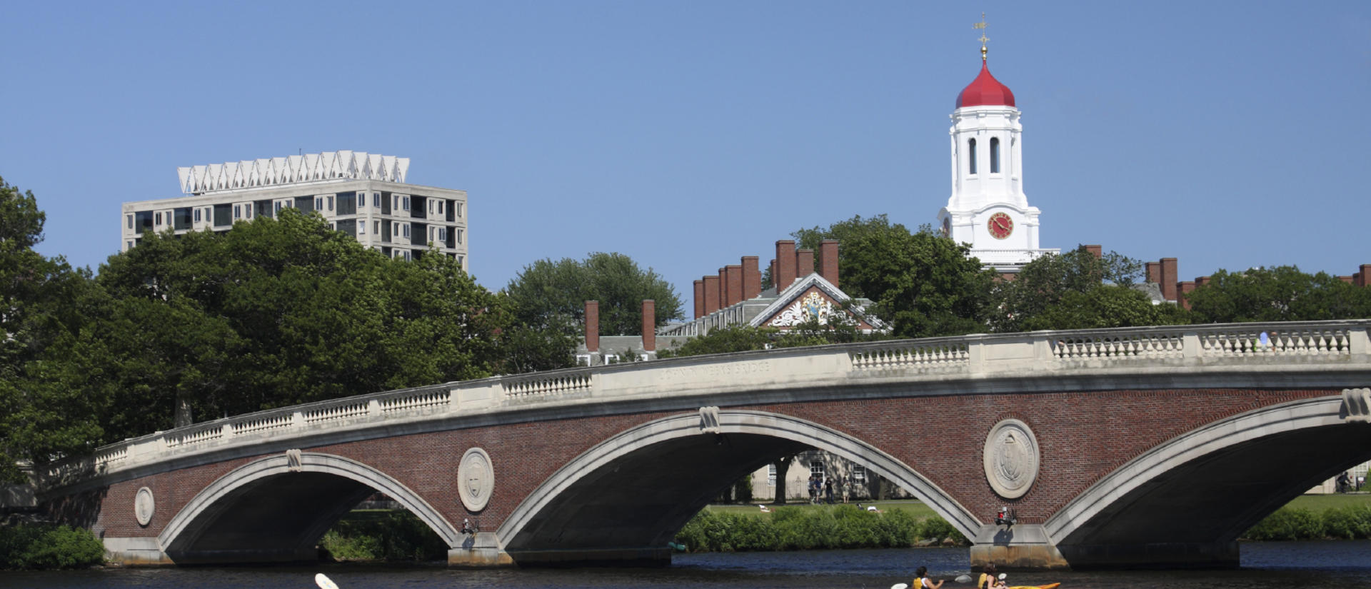 Two rowers pass under a footbridge over the Charles River with Harvard buildings in the background