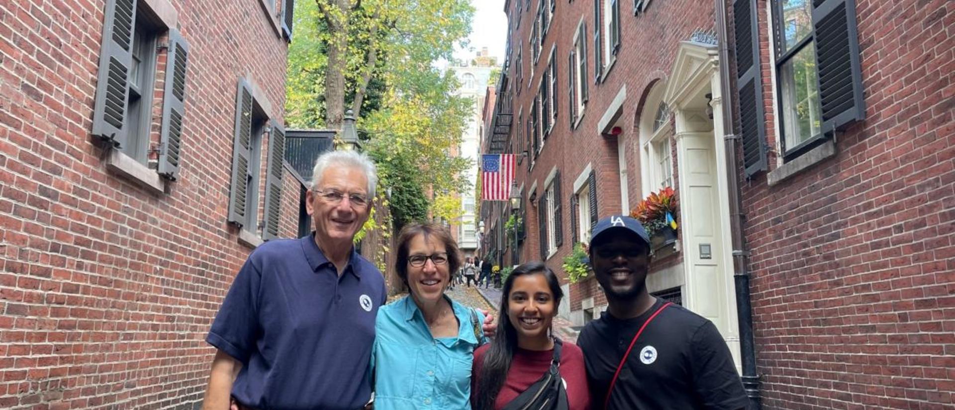 Four people stand shoulder to shoulder between two historic red-brick buildings