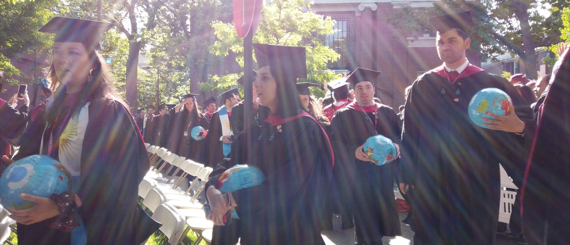 Graduates in caps and gowns process into Harvard Yard under bright sunshine