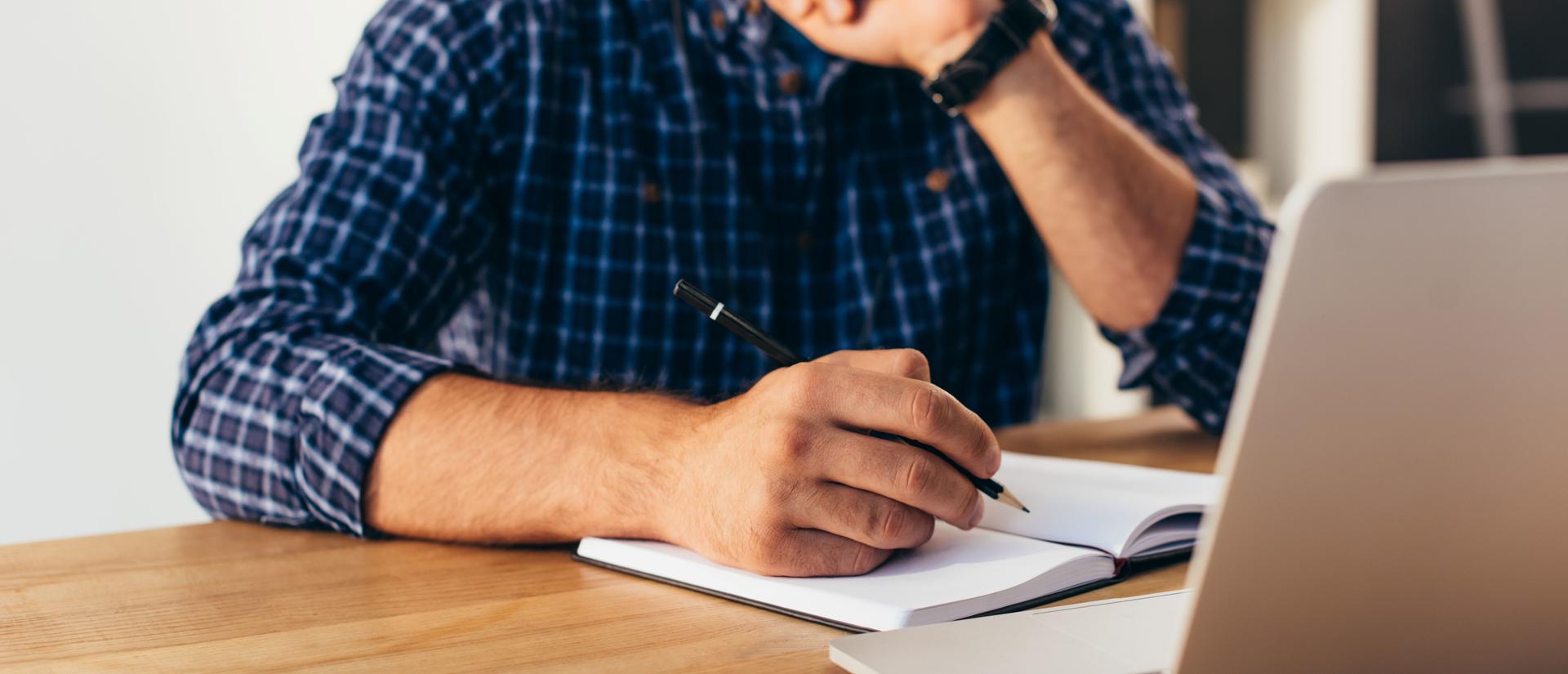 A man takes notes in a notebook while watching a webinar on a laptop