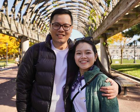 A man stands with his arm around a woman's shoulder under a wooden arch in a public park on a sunny day