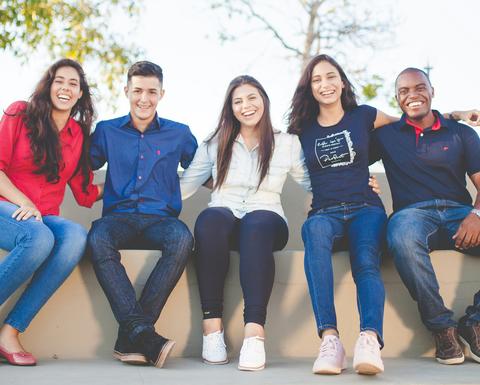 Five smiling students sit on a wall outside