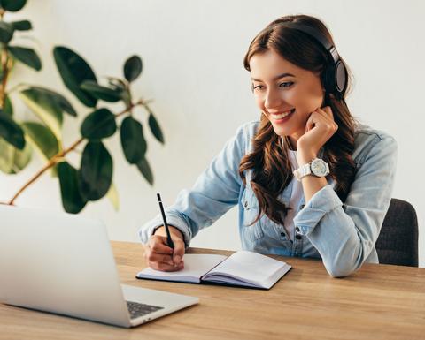 A woman smiles and takes notes while watching a webinar on a laptop